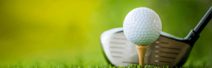 Close up image of a golf club head beside a golf ball on a tee on green grass.